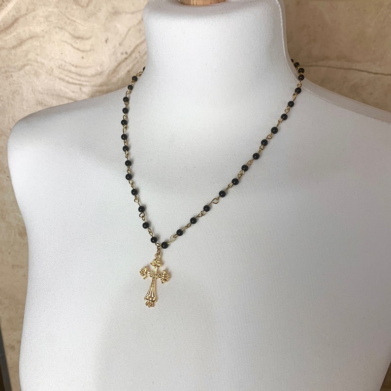 Op de afbeelding: Een zwarte kralenketting met een gouden kruis hanger. Het kruis heeft een gedetailleerd ontwerp.