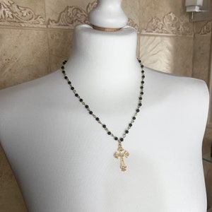 Op de afbeelding: Een zwarte kralenketting met een gouden kruis hanger. Het kruis heeft een gedetailleerd ontwerp.