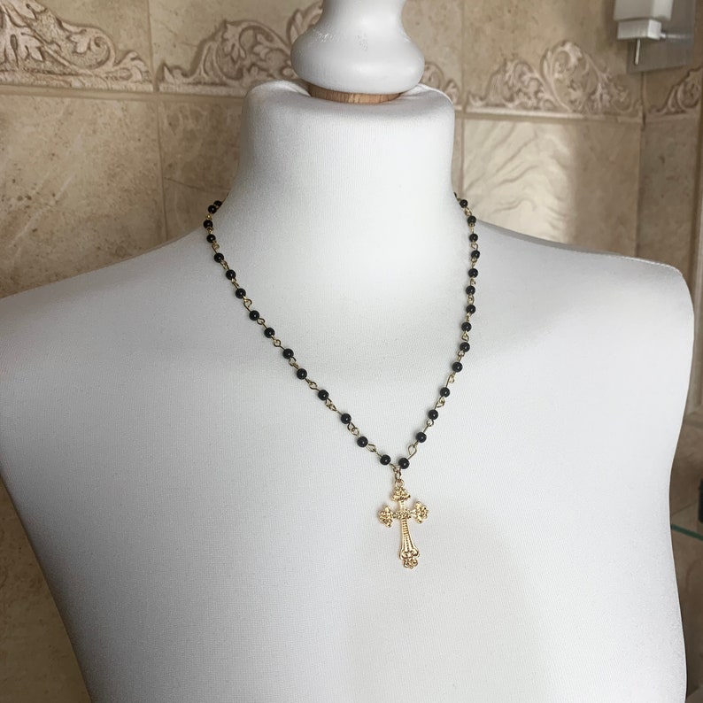 Op de afbeelding: Een zwarte kralenketting met een gouden kruis hanger. Het kruis heeft een gedetailleerd ontwerp.