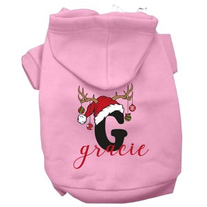 May include: Pink dog hoodie with a Christmas reindeer design featuring a Santa hat, ornaments, and the letter "G" with the name "gracie" written below.