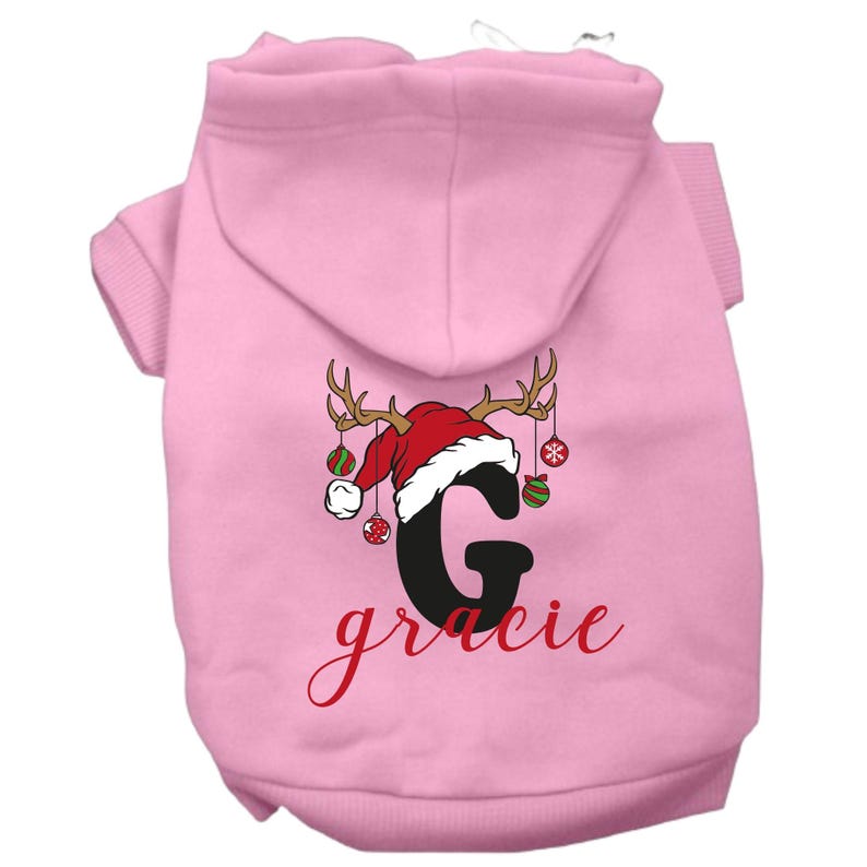 May include: Pink dog hoodie with a Christmas reindeer design featuring a Santa hat, ornaments, and the letter "G" with the name "gracie" written below.