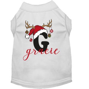 May include: White dog shirt with a red and white Santa hat with antlers and ornaments. The letter G is in black with the name Gracie in red cursive.