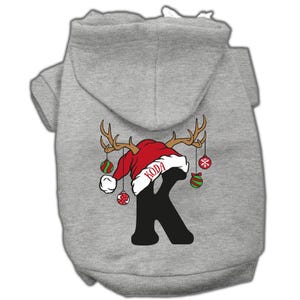 May include: Gray hooded dog sweater with a red Santa hat, reindeer antlers, and a black letter K with the name "Koda" on the hat. The sweater has green and red Christmas ornaments hanging from the antlers.