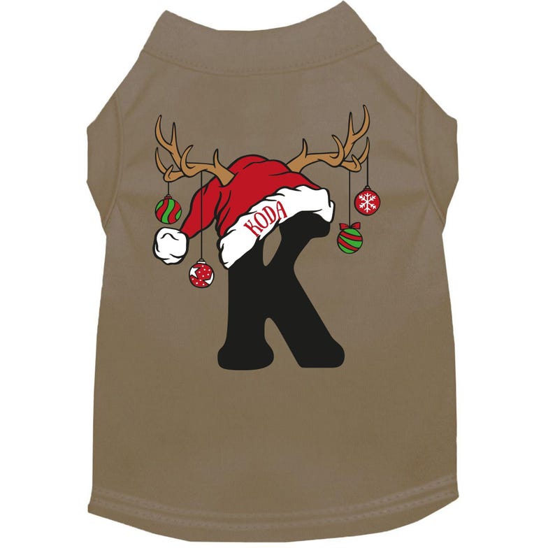 May include: Tan dog shirt with a red Santa hat with antlers, a black letter K, and the name "KODA" on the hat. The shirt has green and red ornaments hanging from the antlers.