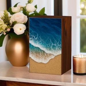 Könnte beinhalten: Eine Holzkiste mit einer Strandszene. Das Bild zeigt blaue Meereswellen, die auf einen sandigen Strand treffen. Die Kiste ist braun mit glänzendem Finish. Links befindet sich eine goldene Vase mit weißen Blumen.