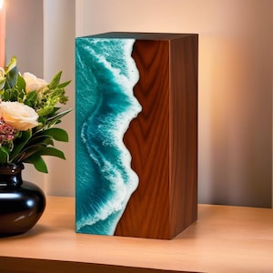 May include: A rectangular wooden box with a striking design. One side features a simulated ocean wave in shades of teal and white resin, contrasting with the warm brown wood grain on the other sides. The box is displayed on a wooden surface.
