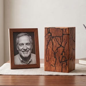 Op de afbeelding: Een houten fotolijst met een zwart-wit foto van een lachende persoon staat naast een houten crematie urn. De urn heeft een uniek, donker, gebarsten ontwerp. De items staan op een houten tafel.