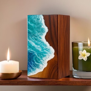 May include: A wooden box with a resin design depicting a beach scene with turquoise water and a sandy shore. A lit candle in a glass jar and a white candle in a brass holder are on a wooden shelf.