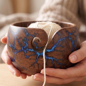 Puede incluir: Un cuenco de hilo de madera con un diseño de resina azul, que contiene una madeja de hilo color crema. El cuenco tiene una ranura curva para pasar el hilo. El cuenco se sostiene con dos manos.