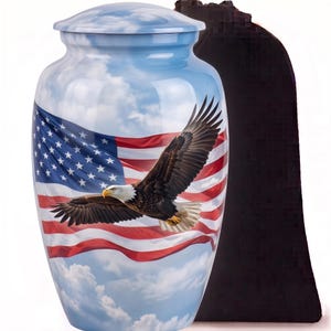May include: A cylindrical cremation urn featuring a glossy finish with a printed design of a bald eagle in flight against a backdrop of a waving American flag and a blue sky with white clouds. The urn has a matching lid and a smooth, rounded shape.