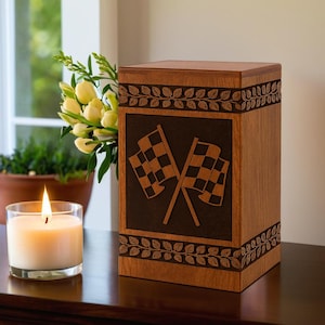May include: A wooden urn with a dark brown square panel featuring crossed checkered flags. The urn is light brown with leaf and vine carvings. A lit candle and flowers are in the background.