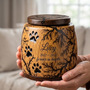 May include: A wooden pet urn with a dark brown lid and base. The urn features paw print cutouts and a tree branch design. The name "Lucy" and dates "2012-2022" are inscribed, along with the phrase "forever in hearts."