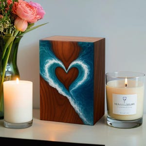 May include: A wooden urn featuring a heart-shaped wave design in blue and white. The urn is made of wood with a natural grain pattern. A lit candle and a vase of flowers are also in the image.