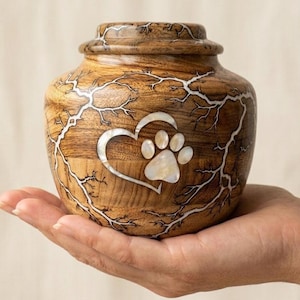 May include: A wooden pet urn with a heart and paw print design. The urn is brown with white accents and a decorative pattern. The heart and paw print are inlaid with a pearlescent material.