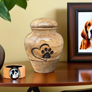 May include: A wooden pet urn with a paw print design inside a heart. The urn is light brown with a lid and sits on a wooden surface. A framed photo of a dog and a small wooden pendant with a dog photo are also present.