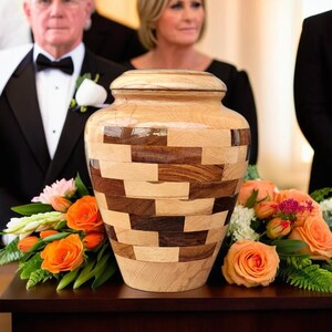 Puede incluir: Una urna de madera con tapa, con un patrón de mosaico de piezas de madera marrón claro y oscuro. La urna se exhibe con rosas naranjas y otras flores, con personas en el fondo.