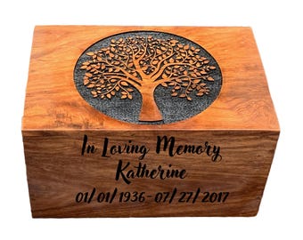 May include: A wooden cremation urn with a carved tree of life design on the lid. The urn is engraved with the text "In Loving Memory Katherine 01/01/1936 - 07/27/2017".