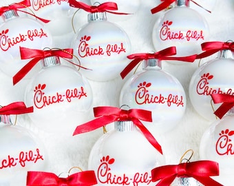 May include: A collection of white, spherical Christmas ornaments, each featuring the red Chick-fil-A logo printed on the front. Every ornament is topped with a silver metal cap and a tied red satin ribbon bow for hanging on a tree.