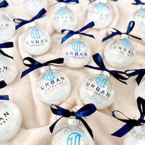 May include: A collection of clear, spherical glass ornaments filled with iridescent material. Each ornament features a dark blue ribbon tied at the top and the printed text "URBAN CATALYST" in blue, accompanied by a matching blue logo icon.