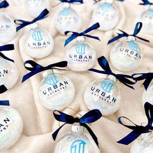 May include: A collection of clear, spherical glass ornaments filled with iridescent white material. Each ornament features a dark blue ribbon tied at the top and the printed text "URBAN CATALYST" in dark blue, accompanied by a matching logo.