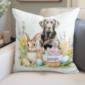 May include: A white throw pillow with a watercolor illustration of a black Labrador Retriever and a brown rabbit. The dog is sitting in a basket filled with colorful Easter eggs. The basket has the word "Bandit" written on it. The pillow has a white background with green, yellow, and blue flowers.