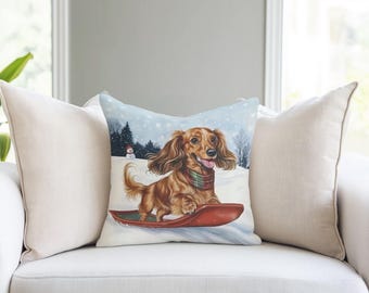 May include: A square throw pillow featuring a long-haired brown dachshund wearing a red and green plaid scarf, riding a red sled through a snowy landscape with a snowman and pine trees in the background, flanked by two plain beige pillows.