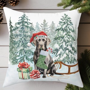May include: A white throw pillow with a winter scene featuring a black Labrador Retriever wearing a Santa hat and holding a candy cane. The dog is standing in front of a snowy forest with a red and green gift box and a sled. The text "DUKE" is on the dog's hat.