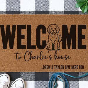 May include: A rectangular doormat with the word "WELCOME" in large, black capital letters, with a cartoon dog illustration and the text "to Charlie's house". The text "...DREW & TAYLOR LIVE HERE TOO" is also included.