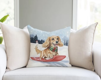 May include: A square throw pillow featuring a light brown long-haired dachshund wearing a red and green striped scarf while riding a red sled on a snowy landscape with a small snowman and evergreen trees in the background.