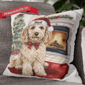 May include: A white throw pillow with a festive Christmas scene featuring a dog wearing a Santa hat. The dog is sitting in front of a fireplace with a Christmas tree in the background. The pillow reads "Hazel".
