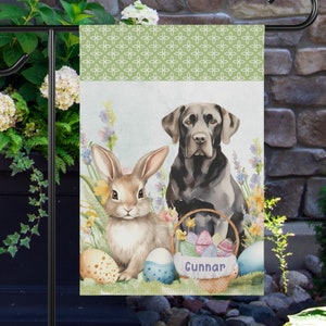 May include: A black Labrador Retriever and a brown bunny rabbit are sitting in a basket filled with colourful Easter eggs. The basket is decorated with a green ribbon and the name "Gunnar" is written on the front. The background is a green and white pattern.