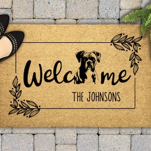 May include: A brown doormat with the text "Welcome the Johnsons" and a black silhouette of a boxer dog. The text is surrounded by a black frame with leafy vines on each side.