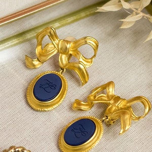May include: A pair of gold tone earrings with blue oval-shaped charms. The charms have a gold rim and a navy blue center with a gold monogram.