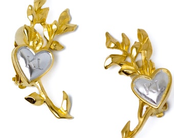 May include: A pair of gold-tone earrings with a heart-shaped pendant featuring the initials "KL" in silver. Each earring has a delicate gold stem with leaves.