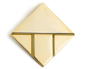 May include: A gold-colored, geometric brooch with a diamond shape. The brooch features a horizontal line and two vertical lines, creating a stylized design. The surface has a polished, reflective finish, and the brooch is set against a white background.