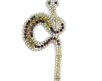 May include: A jeweled snake brooch with a coiled body and a long, straight tail. The brooch features a variety of colored rhinestones, including clear, black, gold, and green, set in a gold-tone metal. The snake's head is at the top, and the tail is at the bottom.