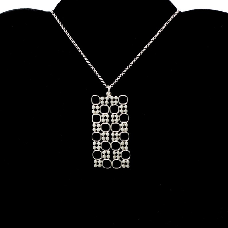 Puede incluir: Un collar plateado con un colgante rectangular. El colgante presenta un dise&ntilde;o de rejilla con elementos cuadrados y circulares. El collar cuelga sobre un fondo negro, resaltando los intrincados detalles de la joya.
