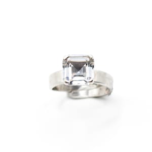May include: A silver-toned ring featuring a large, clear, square-cut gemstone set in a four-prong mounting. The band has a smooth, wide, metallic finish and an open, adjustable design that overlaps slightly at the bottom of the ring structure.