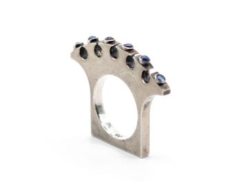 Puede incluir: Un anillo de metal plateado con un borde superior ancho en forma de abanico con seis hendiduras circulares. Cada hendidura contiene una pequeña piedra preciosa azul redonda. El anillo tiene una base rectangular plana y una superficie interior pulida.