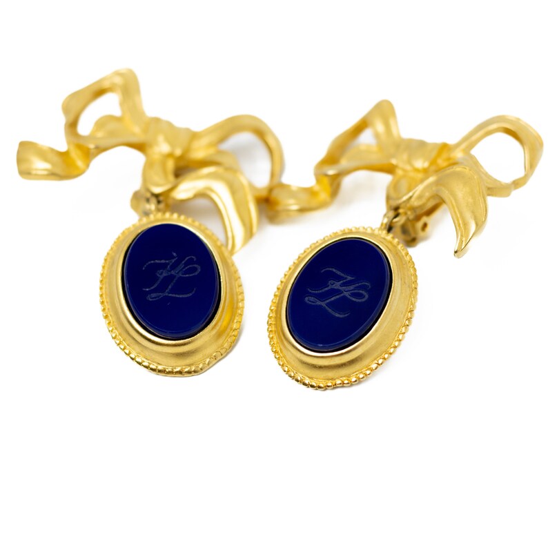 May include: Gold tone earrings with blue oval-shaped stones and a gold bow design. The stones have the initials "ZL" engraved on them.