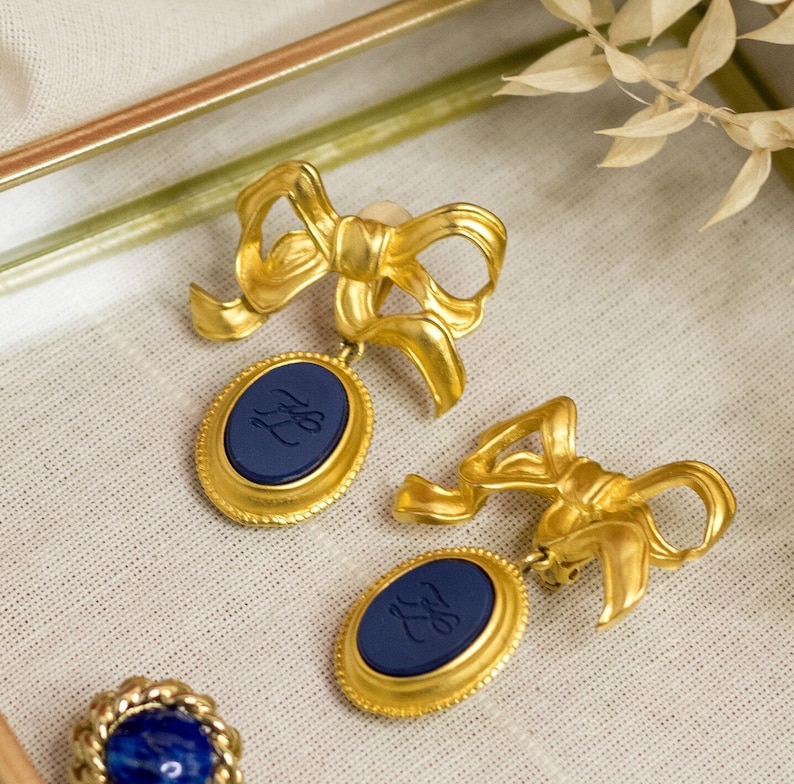 May include: A pair of gold tone earrings with blue oval-shaped charms. The charms have a gold rim and a navy blue center with a gold monogram.