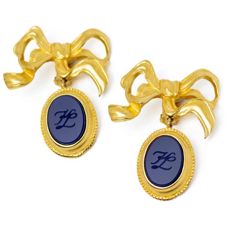 May include: Gold tone earrings with a blue enamel oval pendant featuring the letters "F" and "L" in a cursive script. The earrings are shaped like bows.