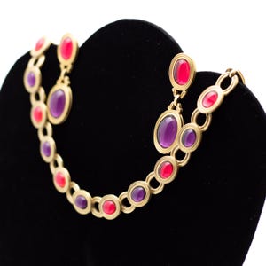 Può includere: Una collana d'oro con pietre ovali di colore viola e rosa alternate. La collana è esposta su un supporto in velluto nero con orecchini abbinati.