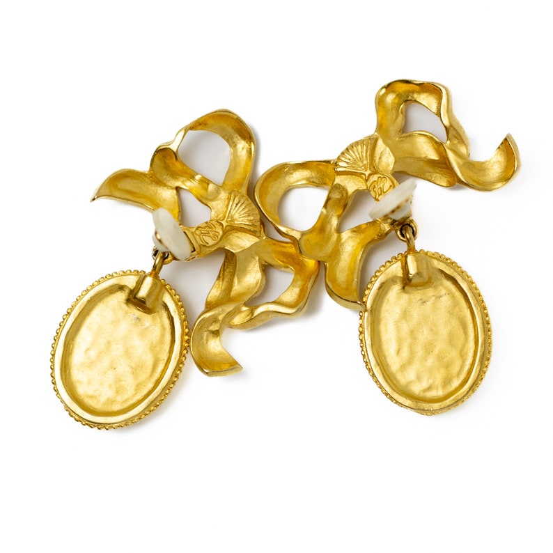 May include: A pair of gold-tone earrings with a bow design and oval-shaped drop pendants. The earrings are made of metal and have a textured finish.