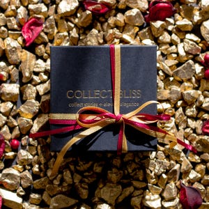 Puede incluir: Una caja de regalo negra con letras doradas y una cinta roja y dorada. La caja est&aacute; rodeada de rocas doradas y p&eacute;talos rojos. El texto de la caja dice "COLLECT BLISS" y "collect stories + elevate elegance."