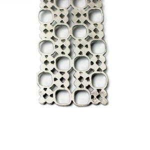 Puede incluir: Primer plano de una pulsera de metal calado en tono plateado. El dise&ntilde;o presenta un patr&oacute;n repetitivo de cuadrados y rombos. La pulsera se muestra sobre un fondo blanco.
