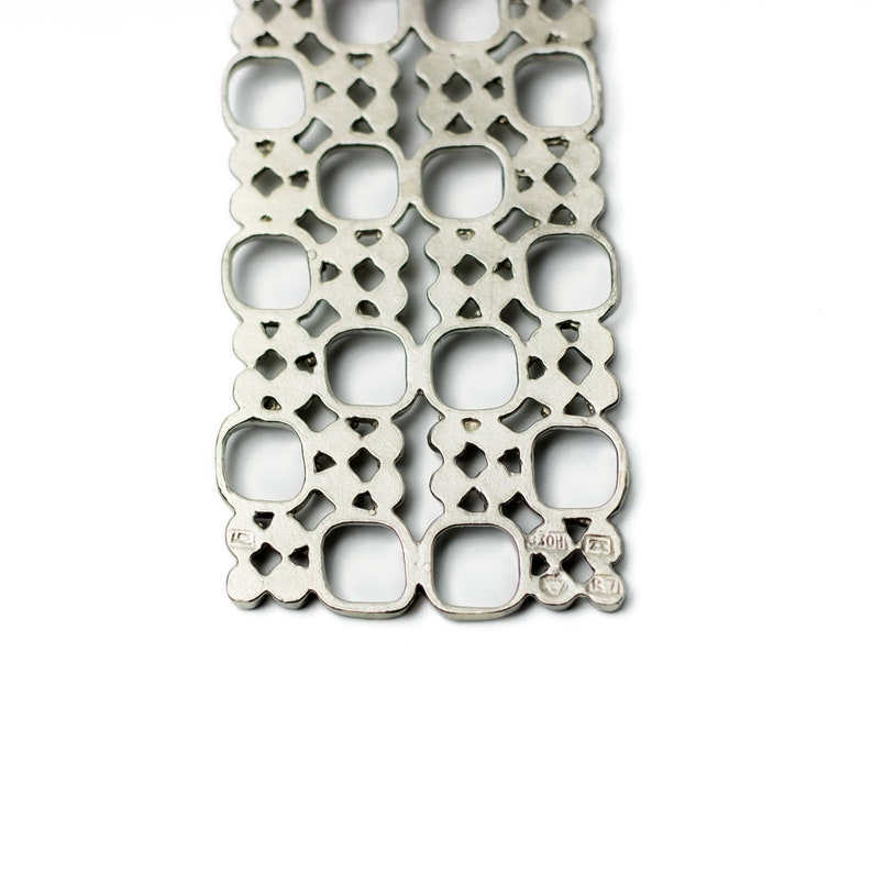 Puede incluir: Primer plano de una pulsera de metal calado en tono plateado. El dise&ntilde;o presenta un patr&oacute;n repetitivo de cuadrados y rombos. La pulsera se muestra sobre un fondo blanco.