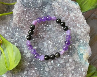 Può includere: Un braccialetto di perline con pietre preziose viola e nere alternate. Il braccialetto è composto da perline nere lucide e rotonde e perline viola sfaccettate. Il braccialetto è esposto su una geode di cristallo viola chiaro.