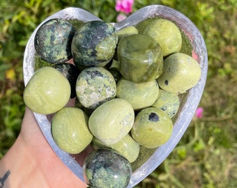May include: A heart-shaped bowl filled with green and black serpentine tumbled stones. The stones are smooth and polished, with a variety of patterns and textures.