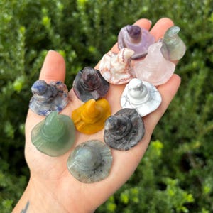 Peut inclure: Plusieurs petites figurines en forme de chapeaux de sorcière, sculptées dans différentes pierres aux couleurs et motifs variés. Les chapeaux, tenus dans une main, présentent des nuances de rose, blanc, jaune et vert, sur fond de feuillage vert.